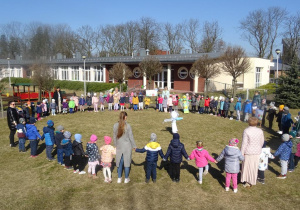 Duża grupa dzieci stoi w kole w ogrodzie przedszkolnym wokół kukły Marzanny.